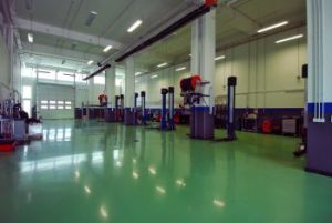Empty industrial workshop with green floor, machinery, overhead pipes, and large windows